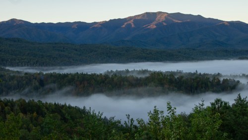 A misty valley surrounded by mountains at dawn, with sunlight illuminating the peaks and trees below.