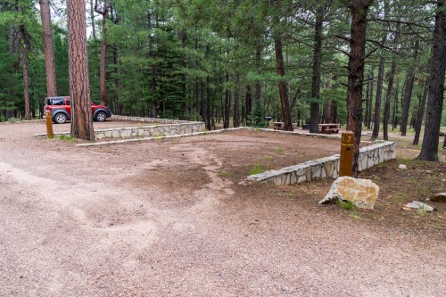 A gravel campsite in a forest with a parked red vehicle, stone borders, and a wooden marker labeled "7."