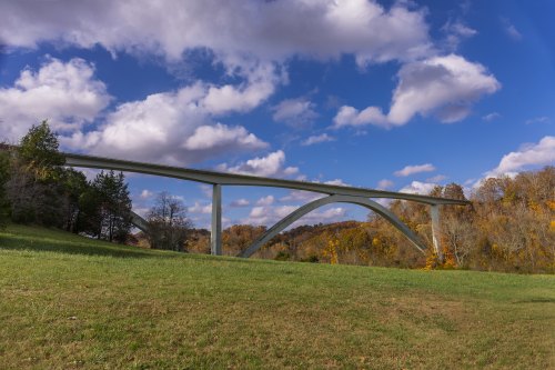 A modern bridge arches over a grassy field, surrounded by trees and colorful autumn foliage under a blue sky.