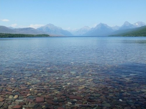 A clear lake with visible colorful stones at the bottom, surrounded by mountains under a blue sky.