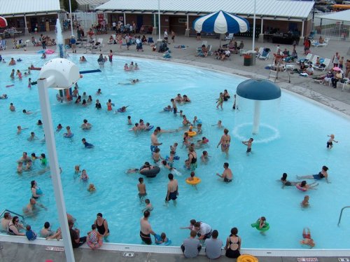 A crowded water park pool filled with people swimming, playing, and relaxing under colorful umbrellas.