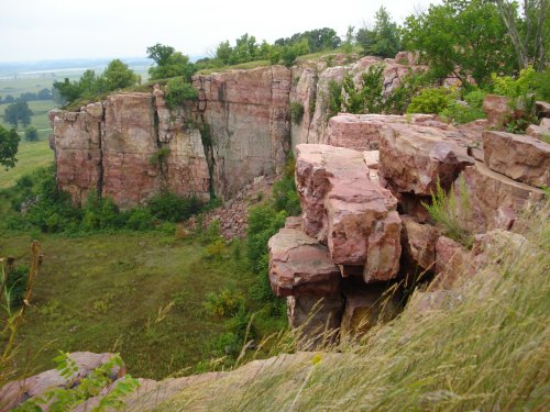Rocky cliffside with layered pink and gray stone, surrounded by green vegetation and a distant landscape.