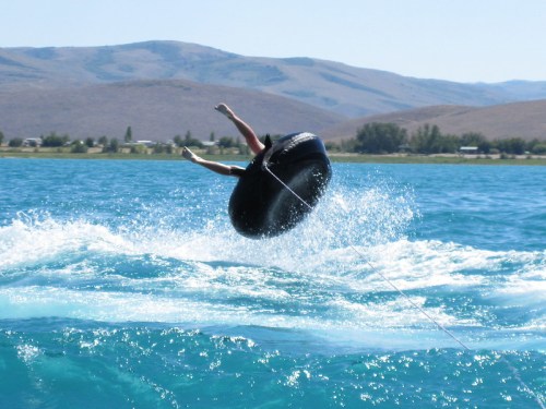 A person is being towed on a black inner tube, flying off into the air above a blue lake with mountains in the background.