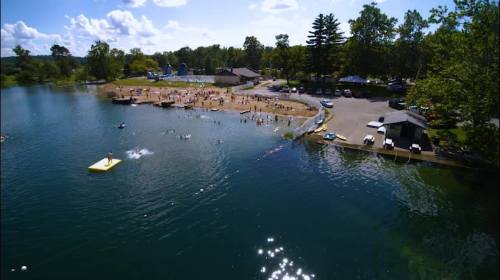 Aerial view of a lake with people swimming and playing, surrounded by trees and a sandy beach area.