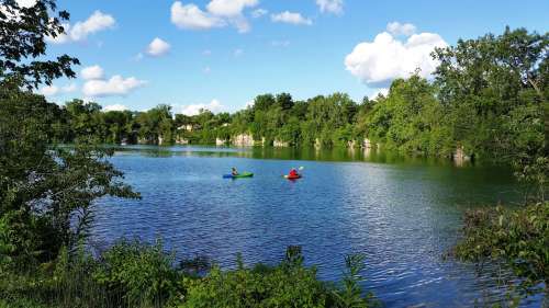 Two kayakers paddle on a calm lake surrounded by lush greenery and blue skies with fluffy clouds.