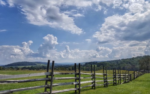 A scenic landscape featuring a wooden fence, green fields, and a blue sky with fluffy clouds and distant hills.