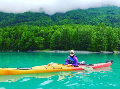 A person in a kayak on turquoise water, surrounded by lush green trees and mountains under a cloudy sky.