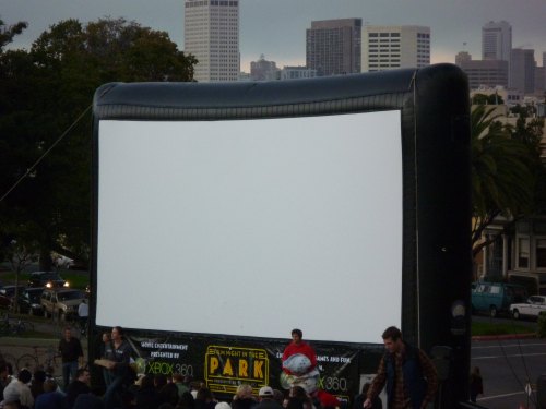 An inflatable outdoor movie screen set up in a park, with people gathered in front, ready for a film screening.