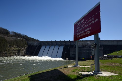 A dam with water flowing through spillways, featuring a warning sign about hazardous conditions.