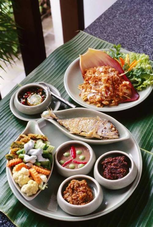 A platter of traditional dishes featuring grilled fish, vegetables, sambal, and a side salad on a banana leaf.