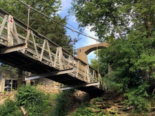 A wooden suspension bridge surrounded by trees, leading to a stone archway in a natural setting.