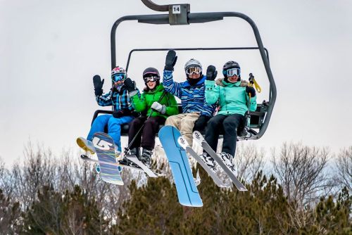 Four skiers on a chairlift, smiling and waving, surrounded by snowy trees and a cloudy sky.