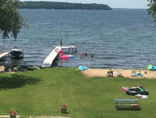 A sunny lakeside scene with people swimming, a dock, and colorful floats on the water, surrounded by green grass.