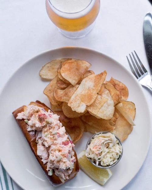 A plate featuring a lobster roll, potato chips, coleslaw, and a lemon wedge, with a beer in the background.