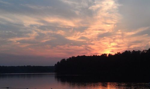 A serene sunset over a calm lake, with colorful clouds reflecting on the water and a silhouette of trees in the foreground.