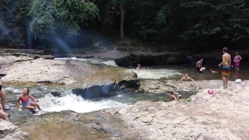 A natural swimming area with people enjoying the water, surrounded by rocks and trees on a sunny day.