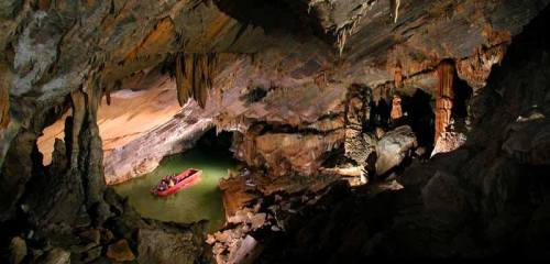 A boat floats on a green lake inside a large, illuminated cave with stalactites and rock formations.