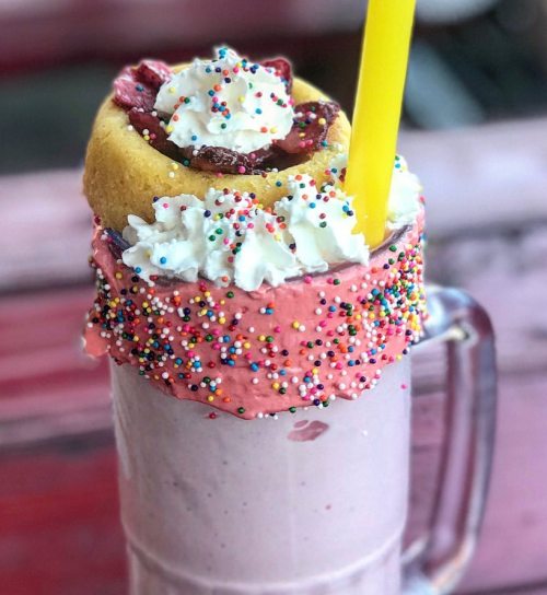 A colorful milkshake topped with whipped cream, sprinkles, and a donut, served with a yellow straw.