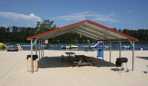 A beach pavilion with picnic tables and a grill, overlooking a sandy area and calm water with inflatable attractions.