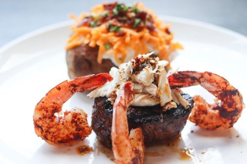 A plate featuring grilled shrimp and a piece of meat topped with shredded crab, alongside a baked potato with cheese.