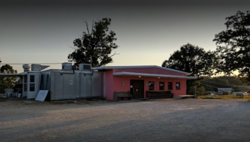 A pink building with a flat roof, surrounded by trees, set against a sunset sky. Gravel path in the foreground.