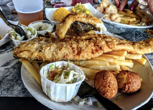 A plate of fried fish, fries, coleslaw, and hushpuppies, with corn and shrimp in the background.