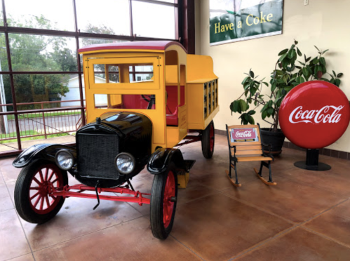 A vintage yellow delivery truck next to a Coca-Cola sign and a small chair, set in a bright indoor space.
