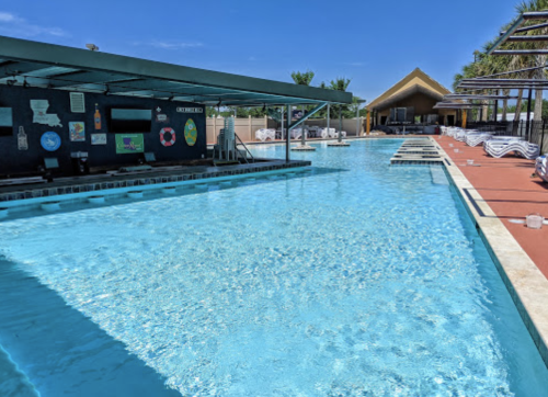 A clear blue swimming pool with lounge chairs and a shaded area in the background under a bright blue sky.
