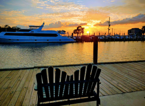 A wooden bench overlooks a marina at sunset, with a yacht and boats silhouetted against a colorful sky.
