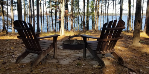 Two wooden chairs facing a fire pit, surrounded by trees near a lake on a sunny day.