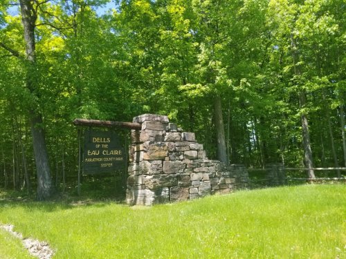 Stone sign for "Dells of the Eau Claire" surrounded by lush green trees and grass.