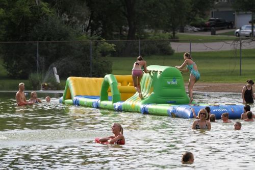 People enjoying a water park inflatable obstacle course in a lake, with swimmers and sunbathers around.