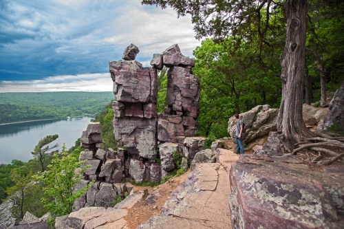 A person stands near towering rock formations overlooking a serene lake surrounded by lush greenery and cloudy skies.