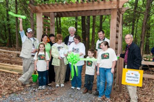 A group of people in matching shirts celebrate a ribbon-cutting ceremony in a wooded area, holding a large green ribbon.
