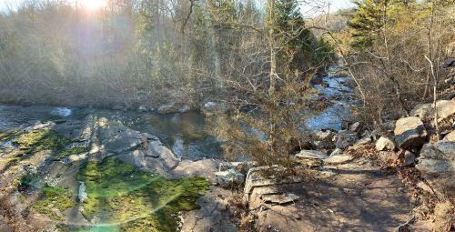 A serene river flows through a rocky landscape, surrounded by trees and sunlight filtering through the branches.