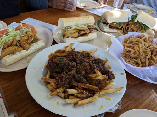 A table with various dishes: a plate of fries topped with beef, two sandwiches, and a basket of onion rings.