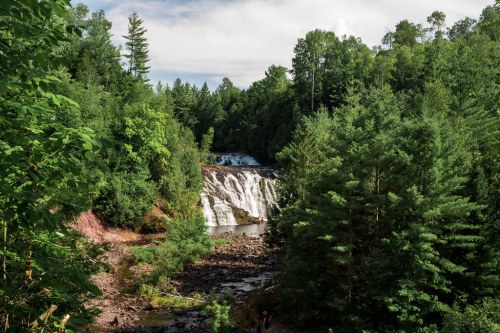 A scenic waterfall surrounded by lush green trees and rocky terrain under a clear blue sky.