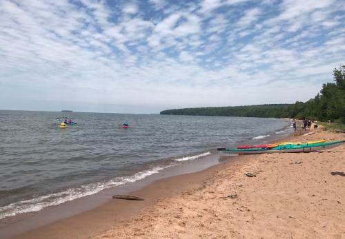 A sandy beach with calm water, kayakers in the distance, and trees lining the shore under a cloudy sky.