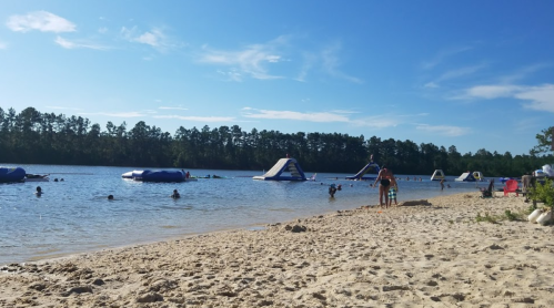 A sandy beach by a lake with people swimming and inflatable structures in the water under a clear blue sky.