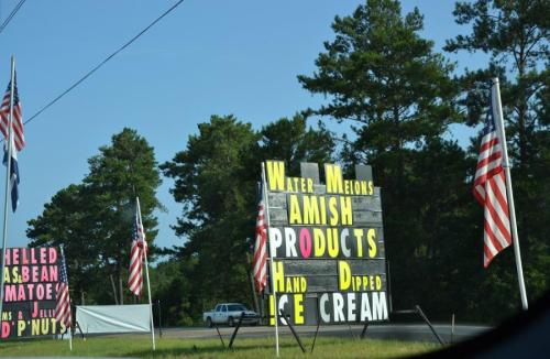 A roadside stand with signs for watermelons, Amish products, and hand-dipped ice cream, surrounded by American flags.
