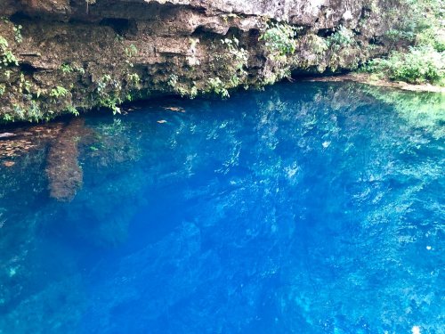 A serene blue water body reflecting rocky cliffs and lush greenery above.