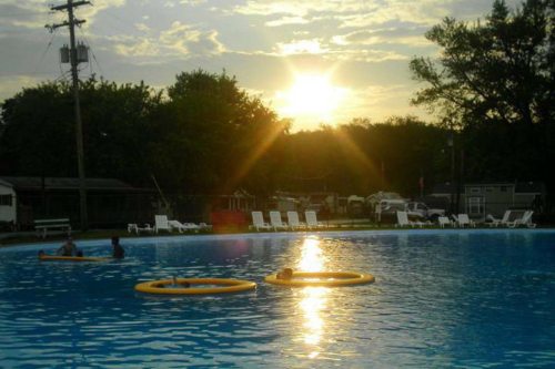 Sunset over a swimming pool with two floating rings and lounge chairs in the background. Trees line the horizon.