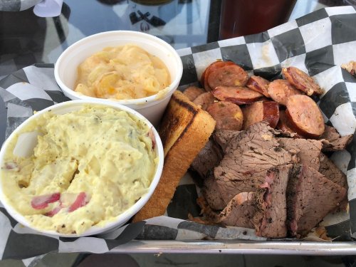 A plate featuring sliced beef, sausage, a slice of toast, and two cups of potato salad and macaroni and cheese.