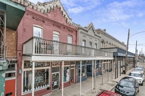 Historic storefronts line a street, featuring colorful facades and a balcony, with parked cars along the curb.