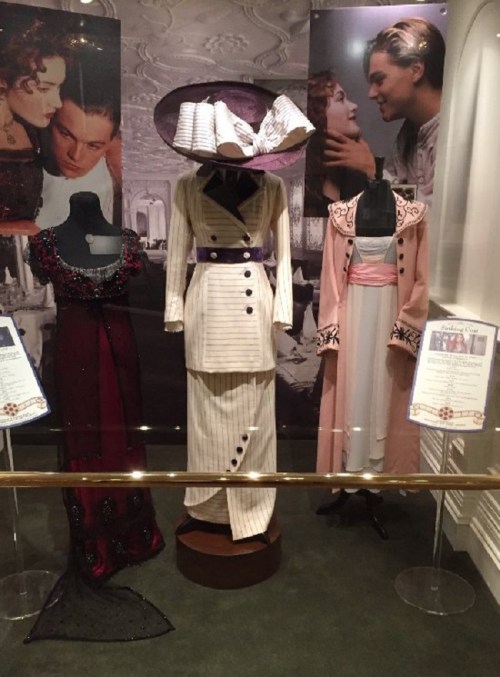Three elegant costumes on mannequins, inspired by early 20th-century fashion, displayed in a museum setting.