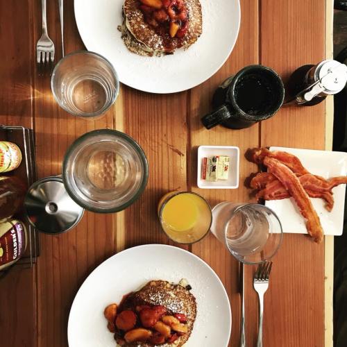 Two plates of pancakes topped with fruit, bacon strips, and drinks on a wooden table.