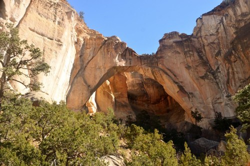 Natural rock arch surrounded by towering cliffs and green vegetation under a clear blue sky.