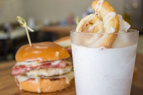 A close-up of a burger with bacon and cheese next to a milkshake topped with a cinnamon roll.