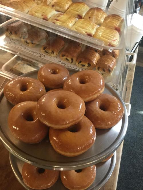 A stack of glazed donuts on a plate, with pastries in a display case in the background.