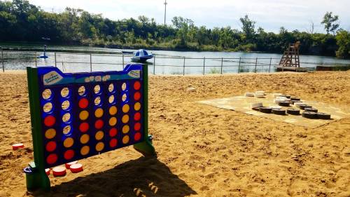 A sandy area by a lake features a large Connect Four game and a checkers board, surrounded by trees and water.